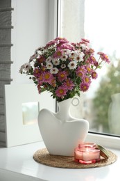 Stylish vase with beautiful flowers and burning candle near window at home