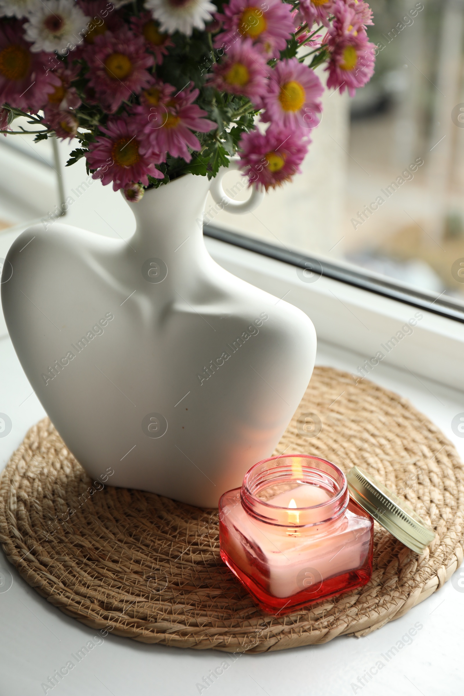 Photo of Stylish vase with beautiful flowers and burning candle near window at home