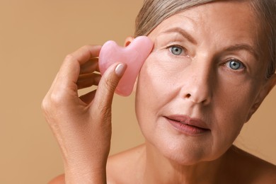 Beautiful woman doing facial massage with gua sha tool on beige background, closeup