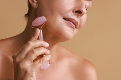 Woman doing facial massage with roller on beige background, closeup