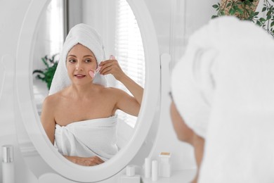 Photo of Smiling woman doing facial self massage with roller near mirror in bathroom