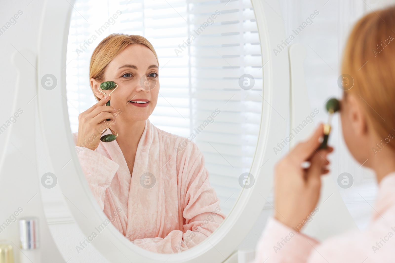 Photo of Smiling woman doing facial self massage with roller near mirror in bathroom