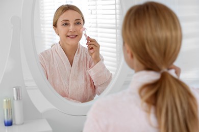 Photo of Smiling woman doing facial self massage with roller near mirror in bathroom