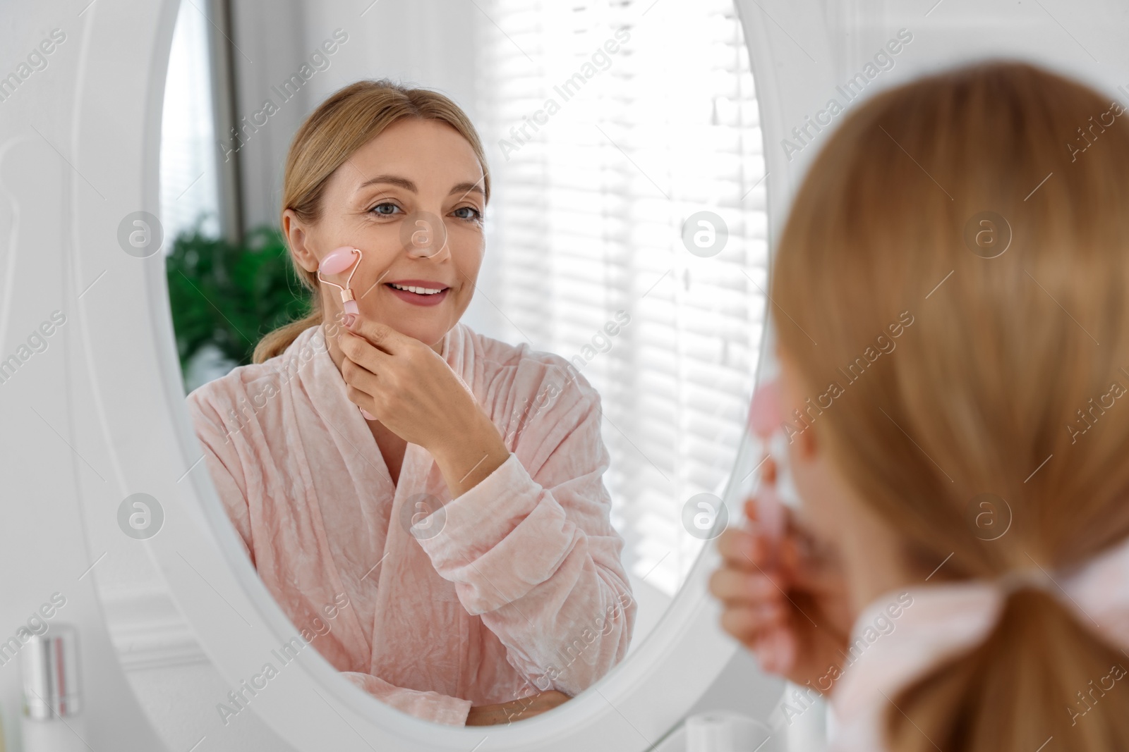 Photo of Smiling woman doing facial self massage with roller near mirror in bathroom