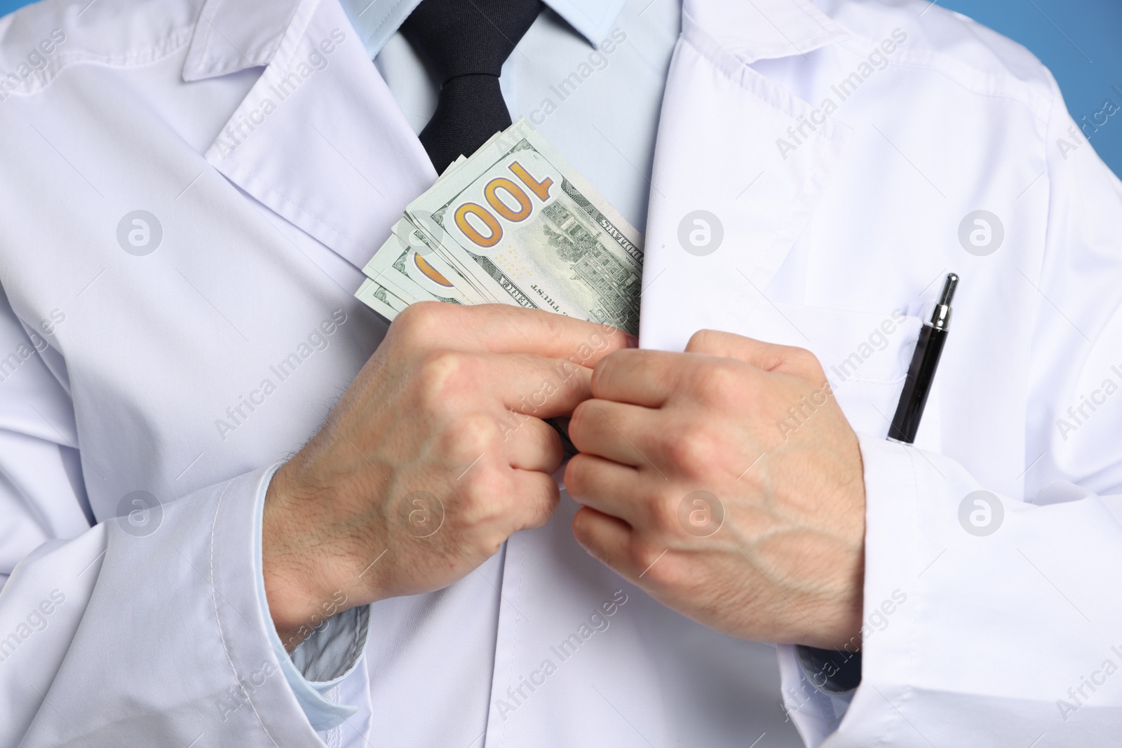 Photo of Corruption concept. Doctor putting dollar banknotes into his coat on blue background, closeup