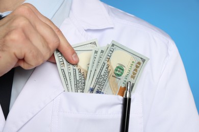 Photo of Corruption concept. Doctor putting dollar banknotes into his pocket on blue background, closeup