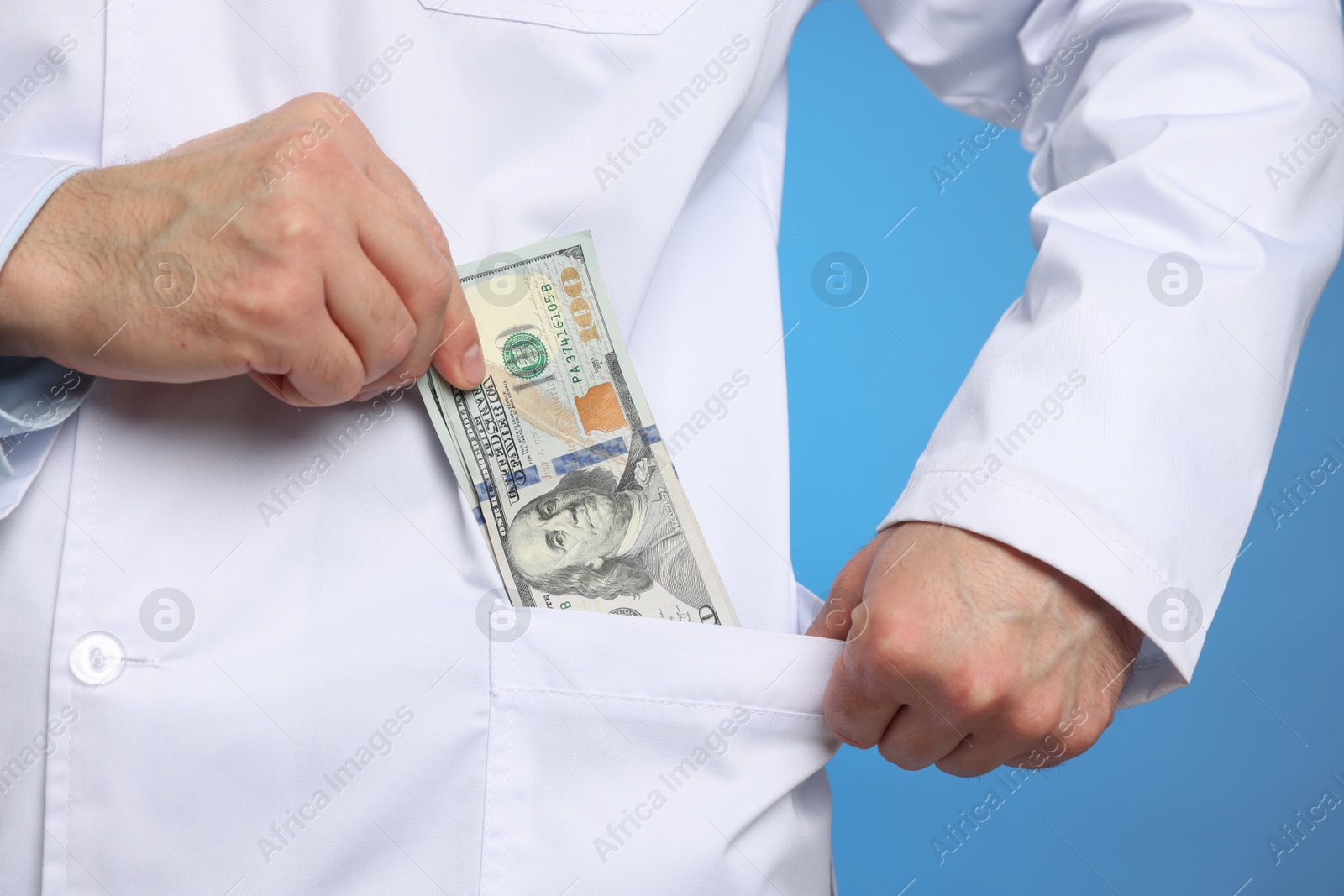 Photo of Corruption concept. Doctor putting dollar banknotes into his pocket on blue background, closeup