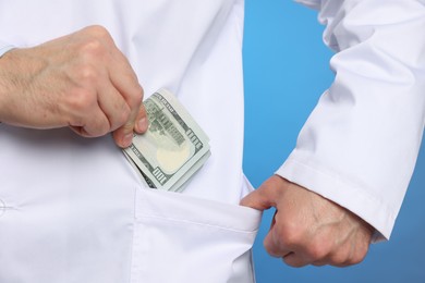 Photo of Corruption concept. Doctor putting dollar banknotes into his pocket on blue background, closeup