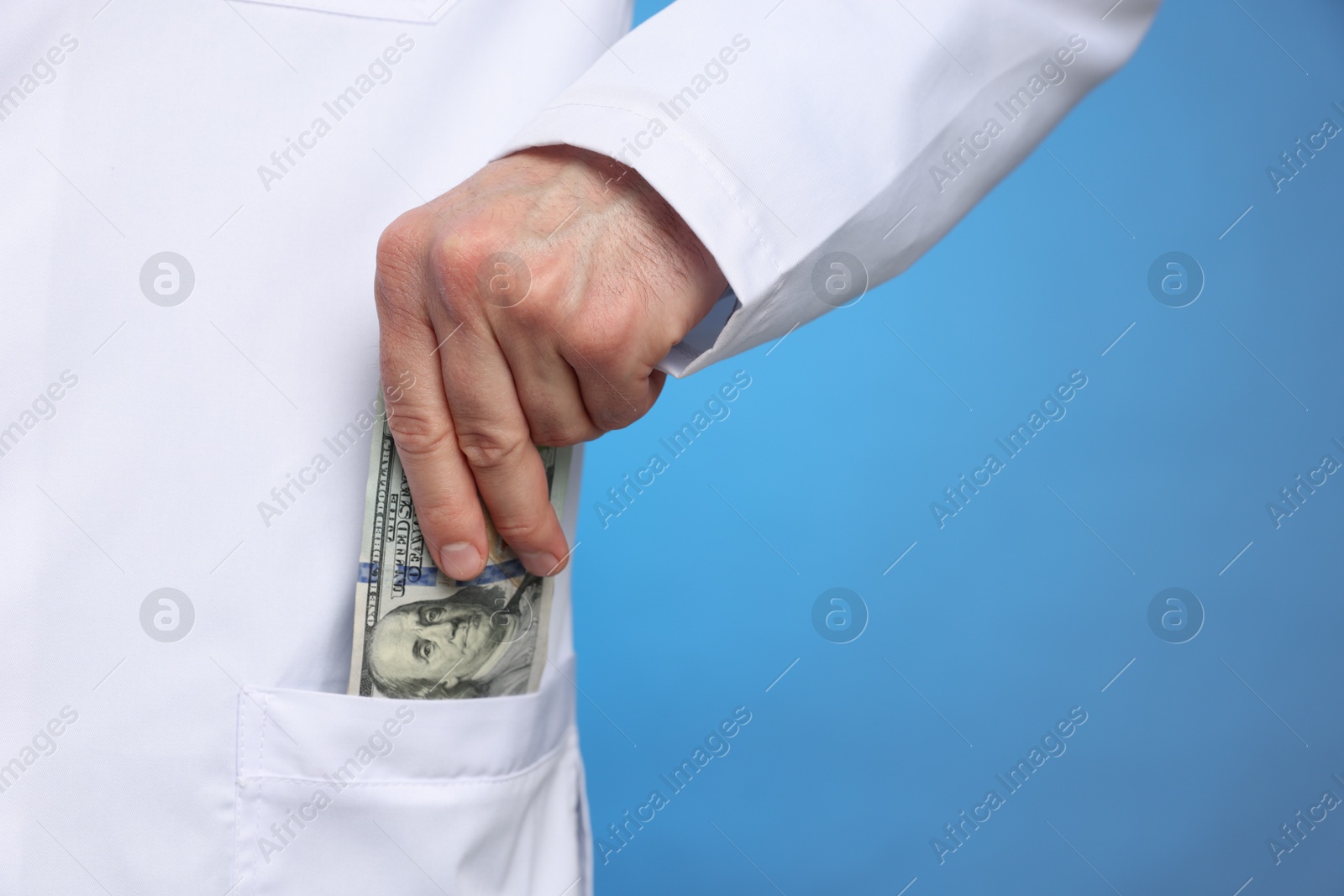 Photo of Corruption concept. Doctor putting dollar banknotes into his pocket on blue background, closeup. Space for text