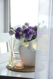 Photo of Stylish vase with beautiful flowers, candle and reed diffuser near window at home