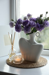 Stylish vase with beautiful flowers, candle and reed diffuser near window at home