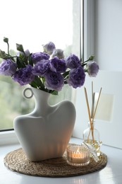 Stylish vase with beautiful flowers, candle and reed diffuser near window at home