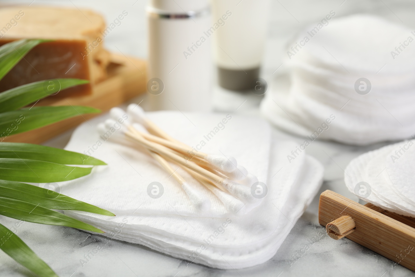 Photo of Cotton pads, swabs cosmetic products and palm leaf on light marble table, closeup