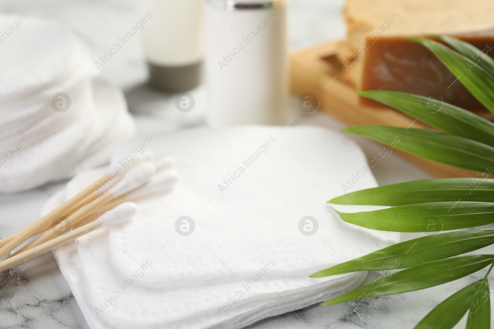 Photo of Cotton pads, swabs cosmetic products and palm leaf on light marble table, closeup