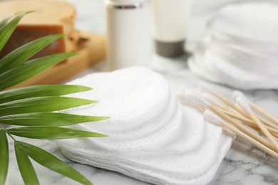 Photo of Cotton pads, swabs cosmetic products and palm leaf on light marble table, closeup