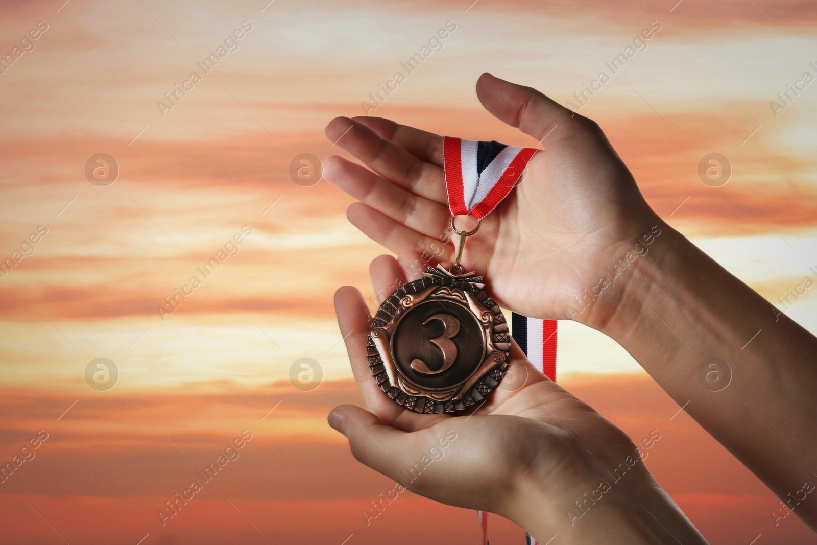 Image of Woman holding bronze medal in hand against sunset sky, closeup