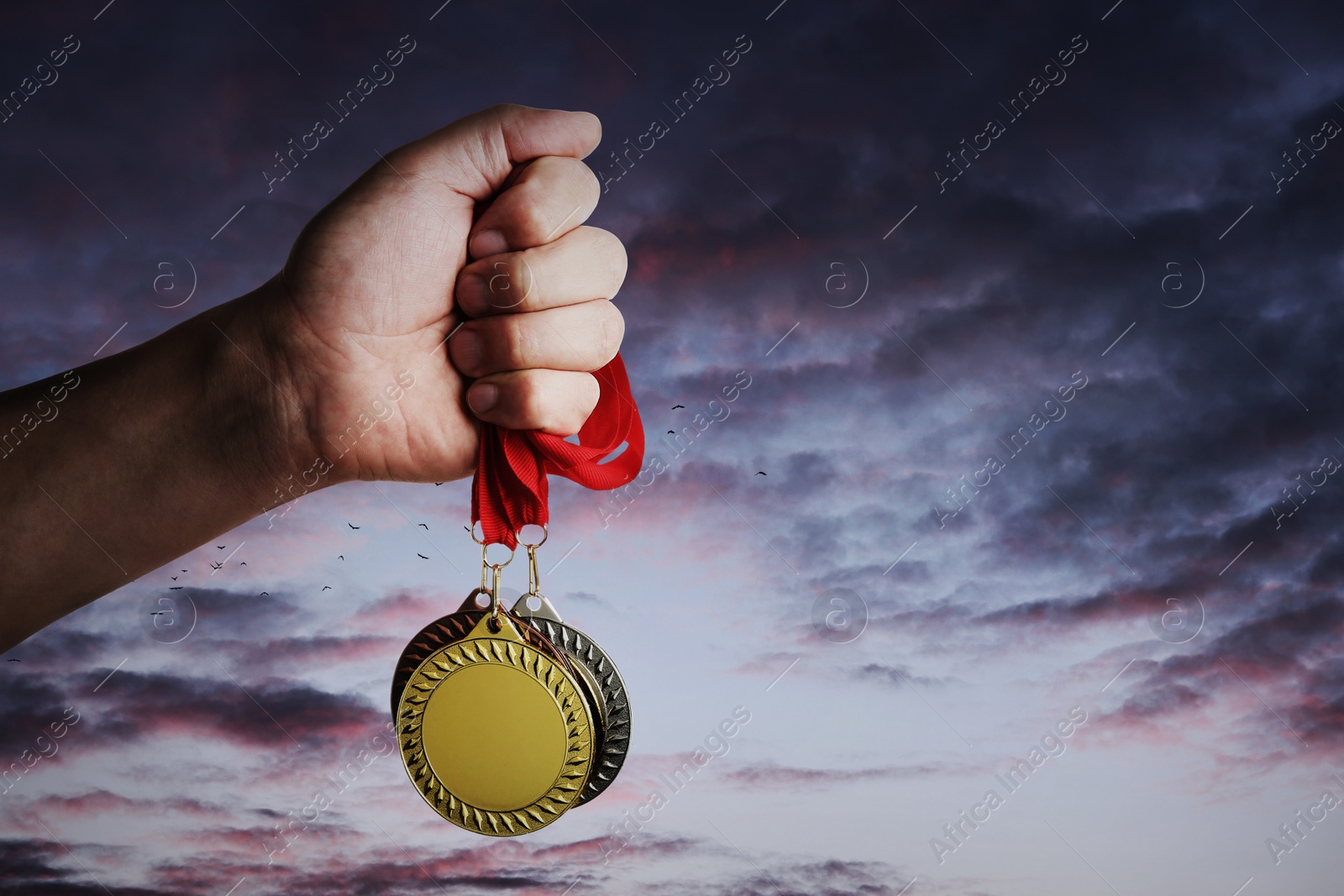 Image of Man holding gold, silver and bronze medals in hand against sunset sky, closeup