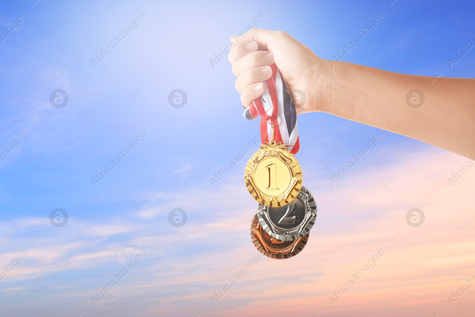 Image of Woman holding gold, silver and bronze medals in hand against sky, closeup