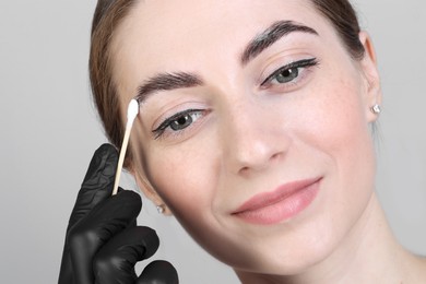 Photo of Brow lamination. Cosmetologist applying cream onto woman's eyebrows against grey background, closeup