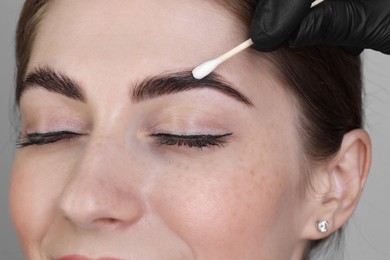 Photo of Brow lamination. Cosmetologist applying cream onto woman's eyebrows against grey background, closeup