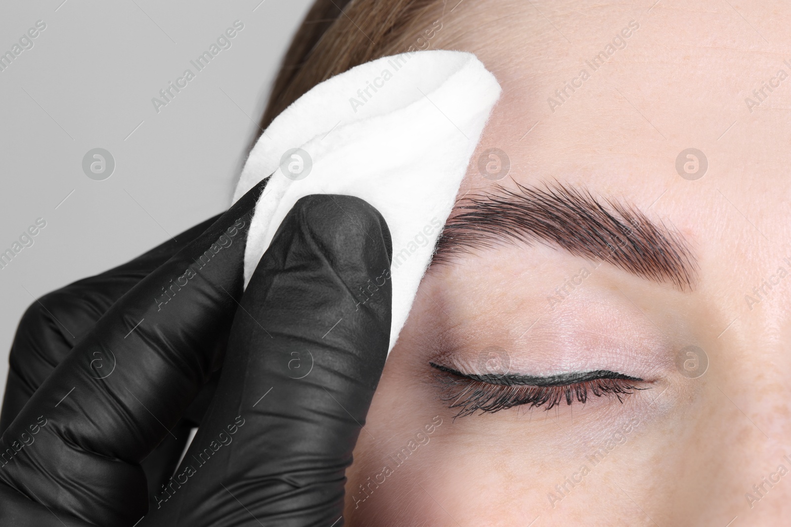 Photo of Brow lamination. Cosmetologist wiping woman's eyebrows with cotton pad against grey background, closeup