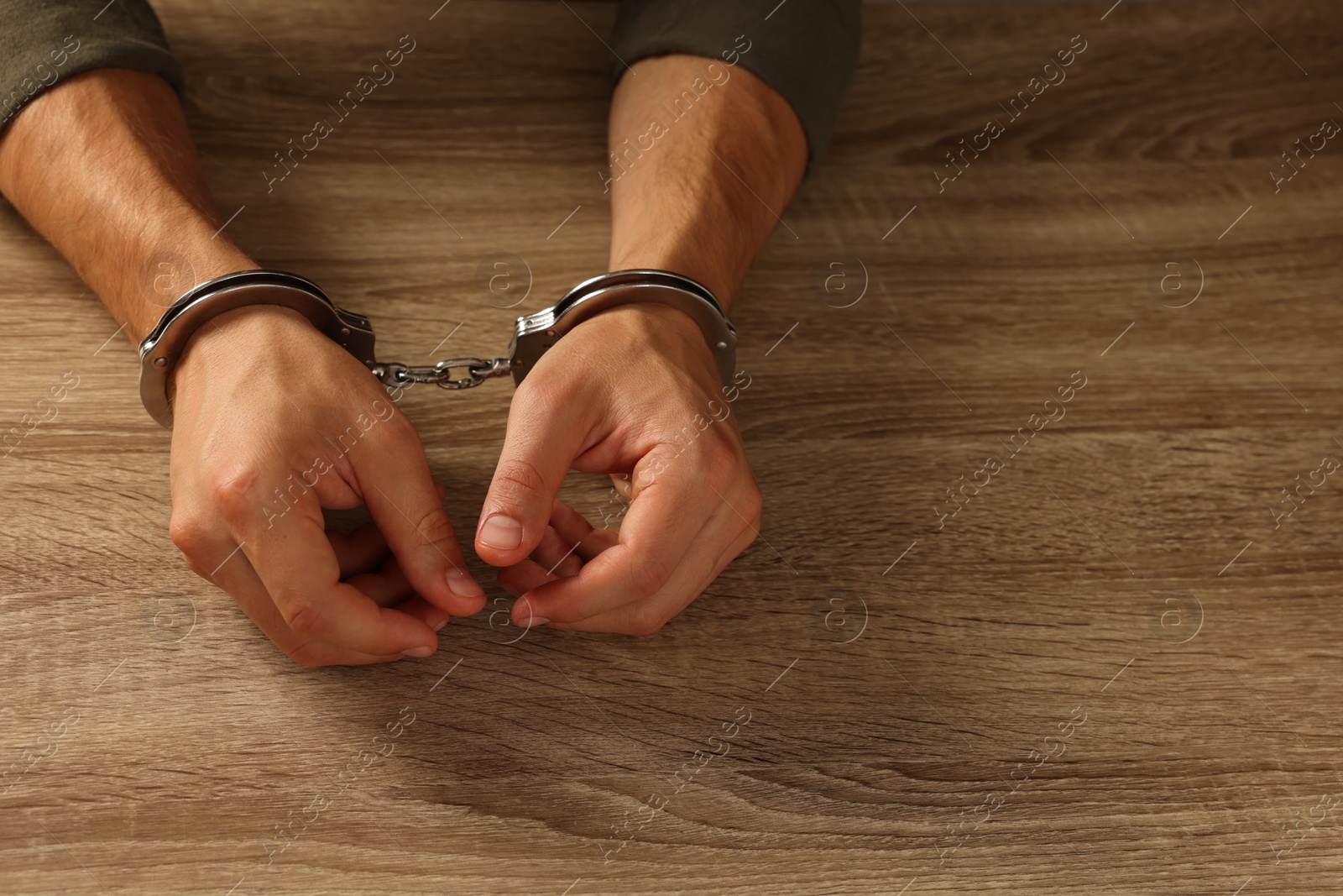 Photo of Man in metal handcuffs at wooden table, closeup. Space for text