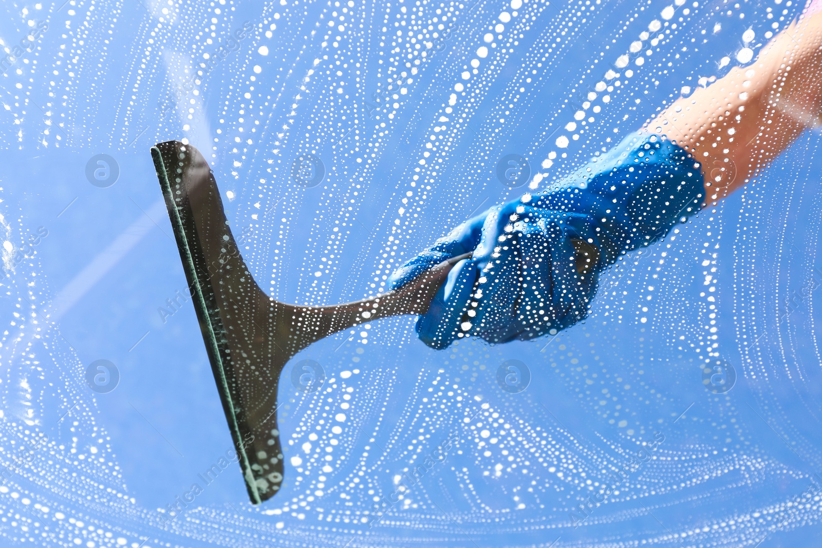 Photo of Woman washing window with squeegee tool against blue sky, closeup