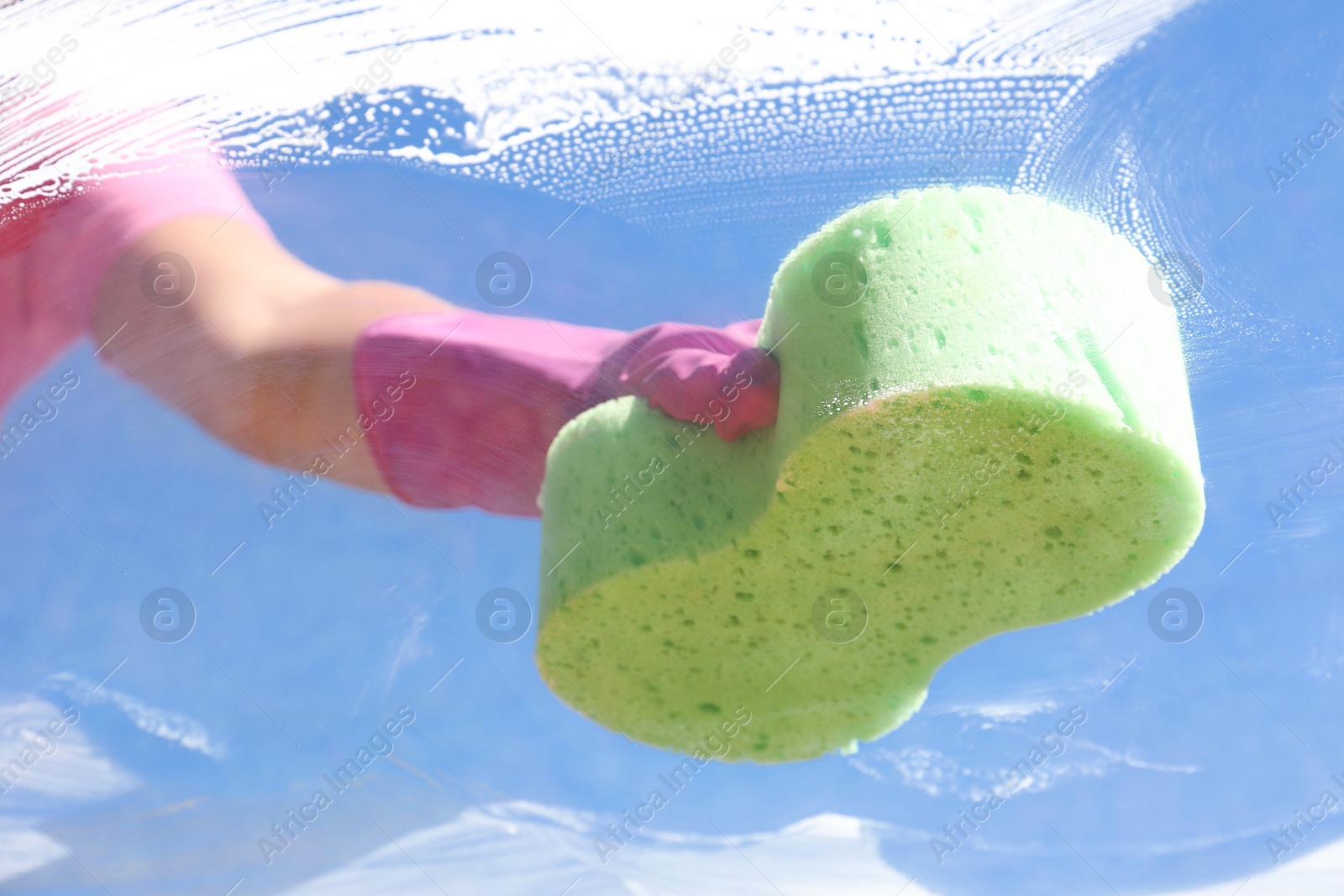 Photo of Woman washing window with sponge against blue sky, closeup