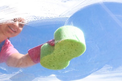 Photo of Woman washing window with sponge against blue sky, closeup