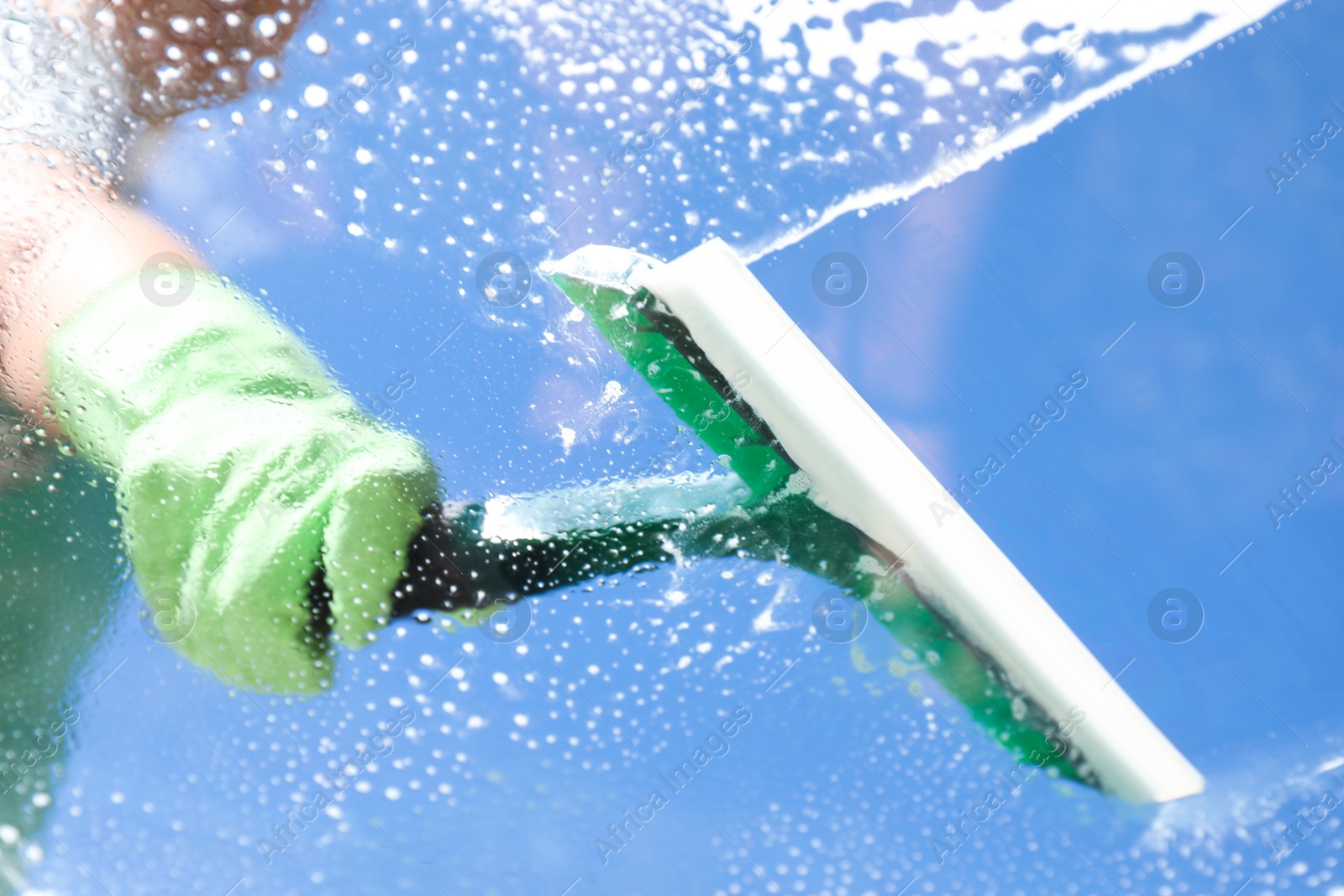 Photo of Woman washing window with squeegee tool against blue sky, closeup