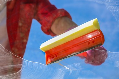 Photo of Woman washing window with squeegee tool against blue sky, closeup