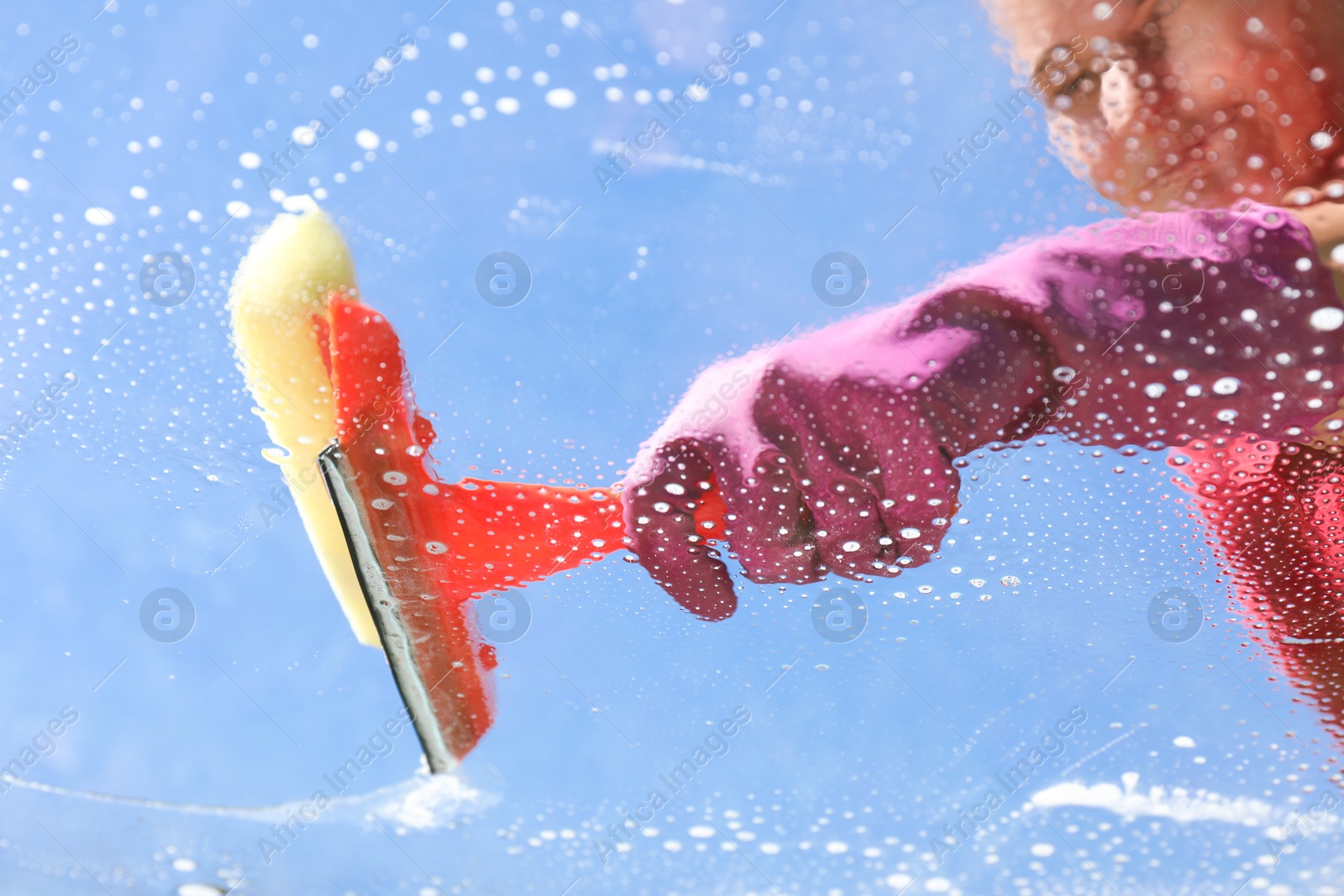 Photo of Woman washing window with squeegee tool against blue sky, closeup