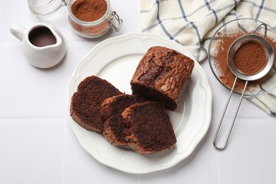 Photo of Delicious cut chocolate sponge cake and ingredients on white tiled table, flat lay