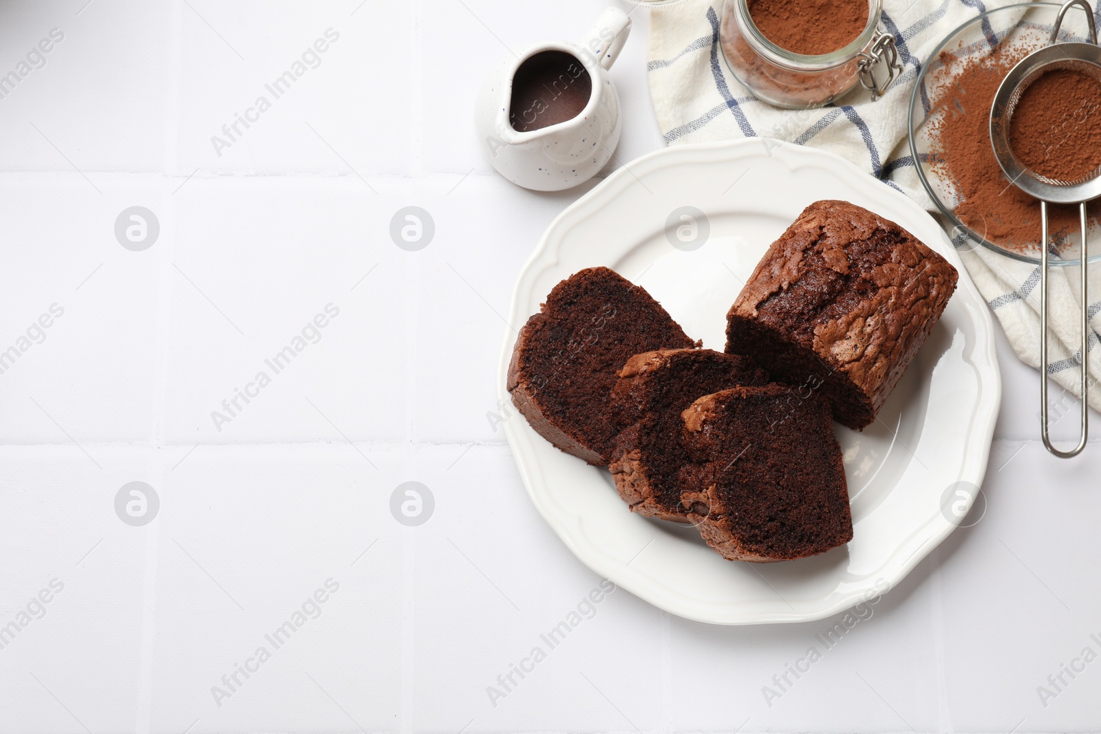 Photo of Delicious cut chocolate sponge cake and ingredients on white tiled table, flat lay. Space for text