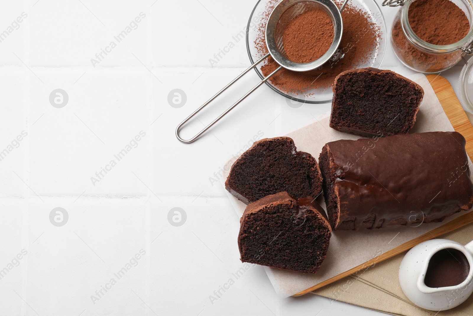 Photo of Delicious cut chocolate sponge cake and ingredients on white tiled table, flat lay. Space for text