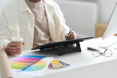 Designer with paper cup of drink working on tablet at table in office, closeup