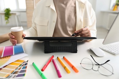 Designer with paper cup of drink using tablet at table in office, closeup