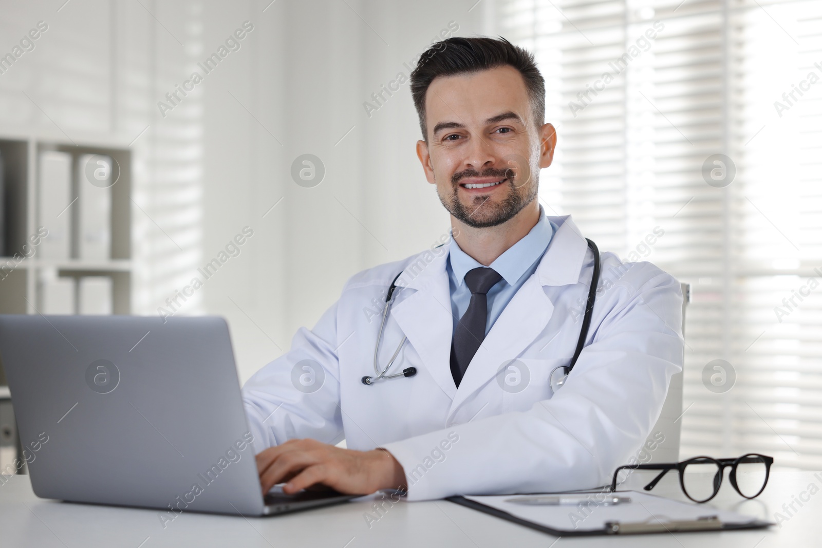 Photo of Smiling doctor working with laptop at table in clinic