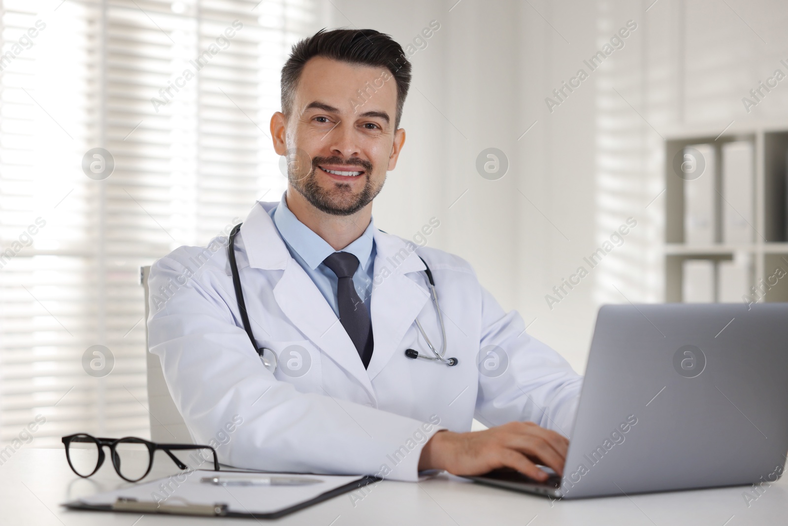 Photo of Smiling doctor working with laptop at table in clinic