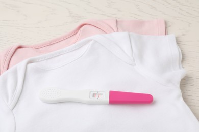 Pregnancy test and baby onesies on white wooden table, above view