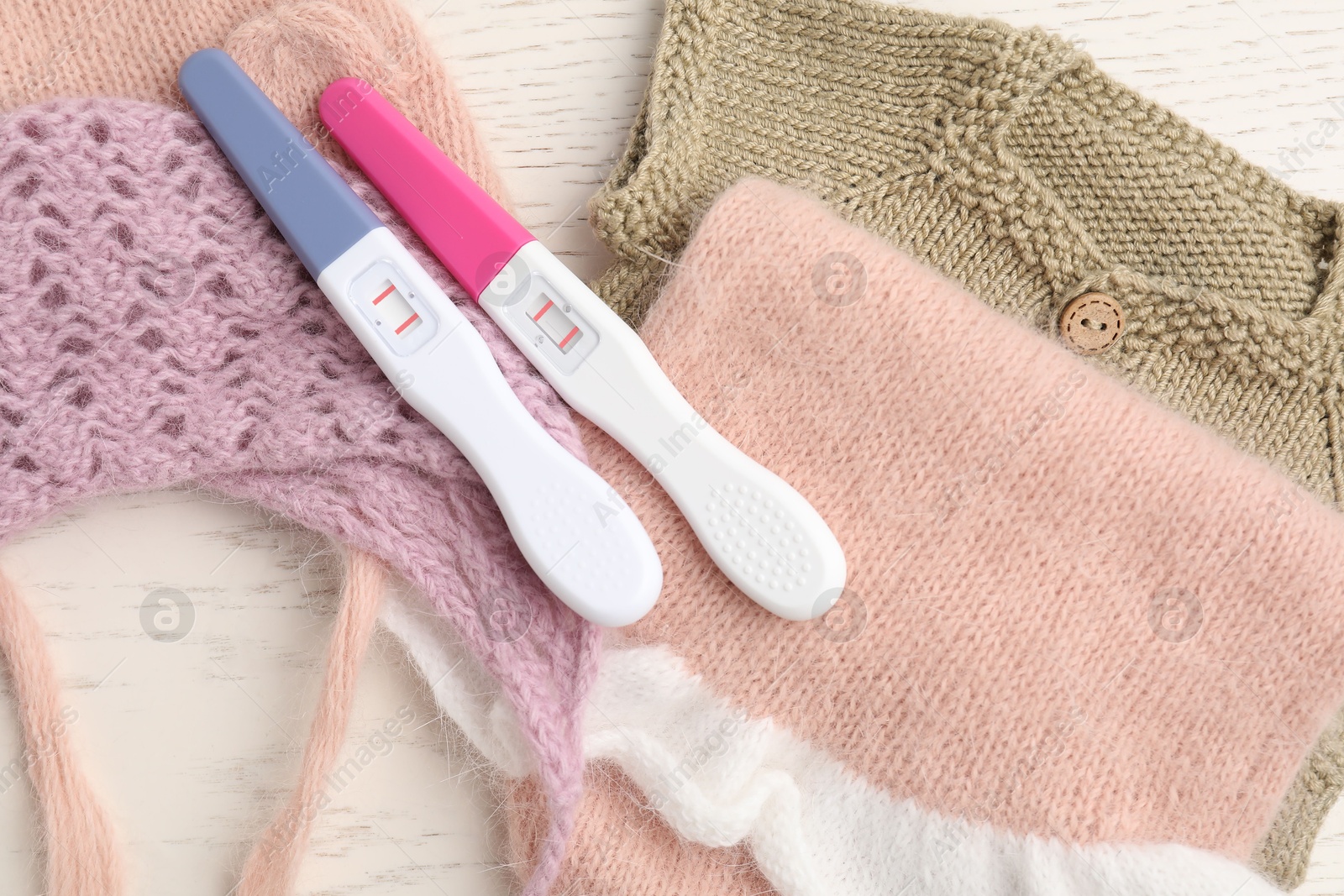 Photo of Two pregnancy tests and baby clothes on white wooden table, flat lay