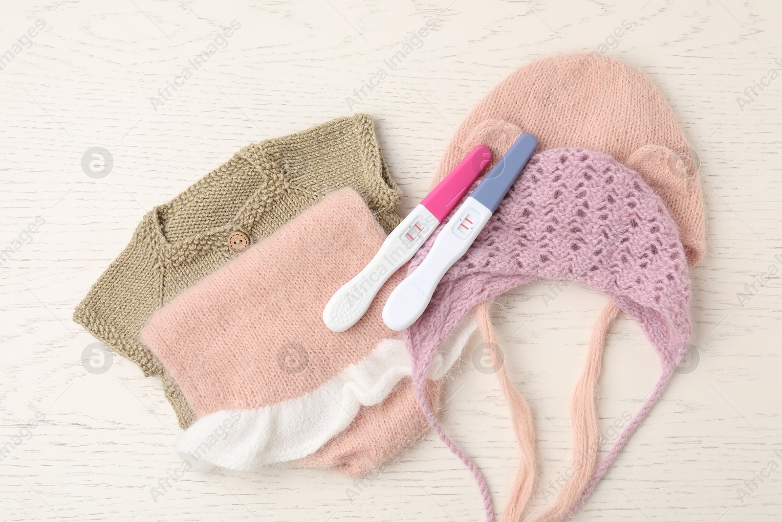 Photo of Two pregnancy tests and baby clothes on white wooden table, flat lay