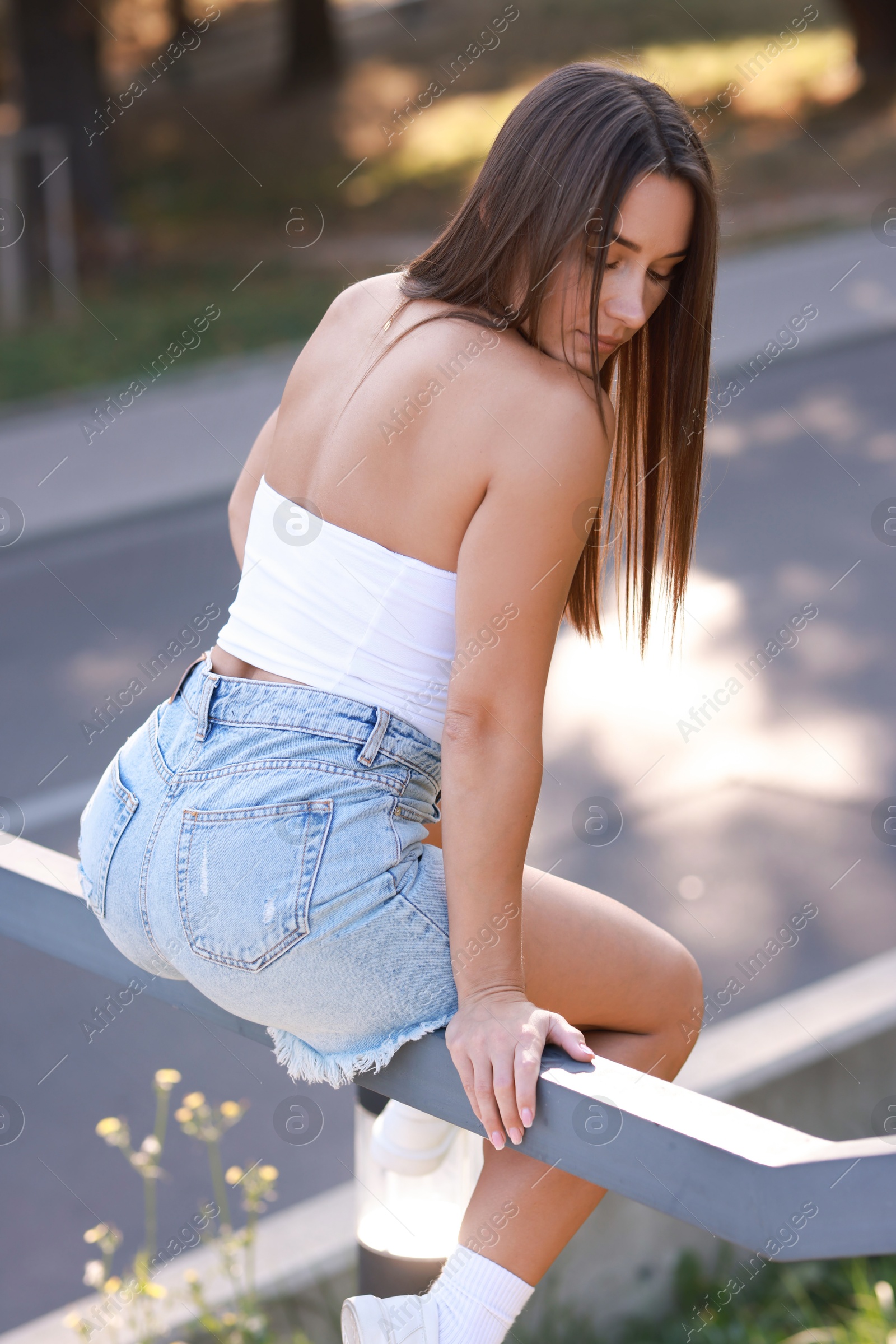 Photo of Beautiful woman in stylish denim shorts sitting on metal handrail outdoors