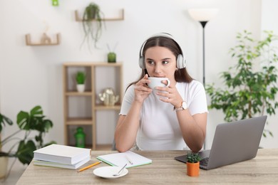 Student in headphones enjoying hot drink while studying at wooden table indoors