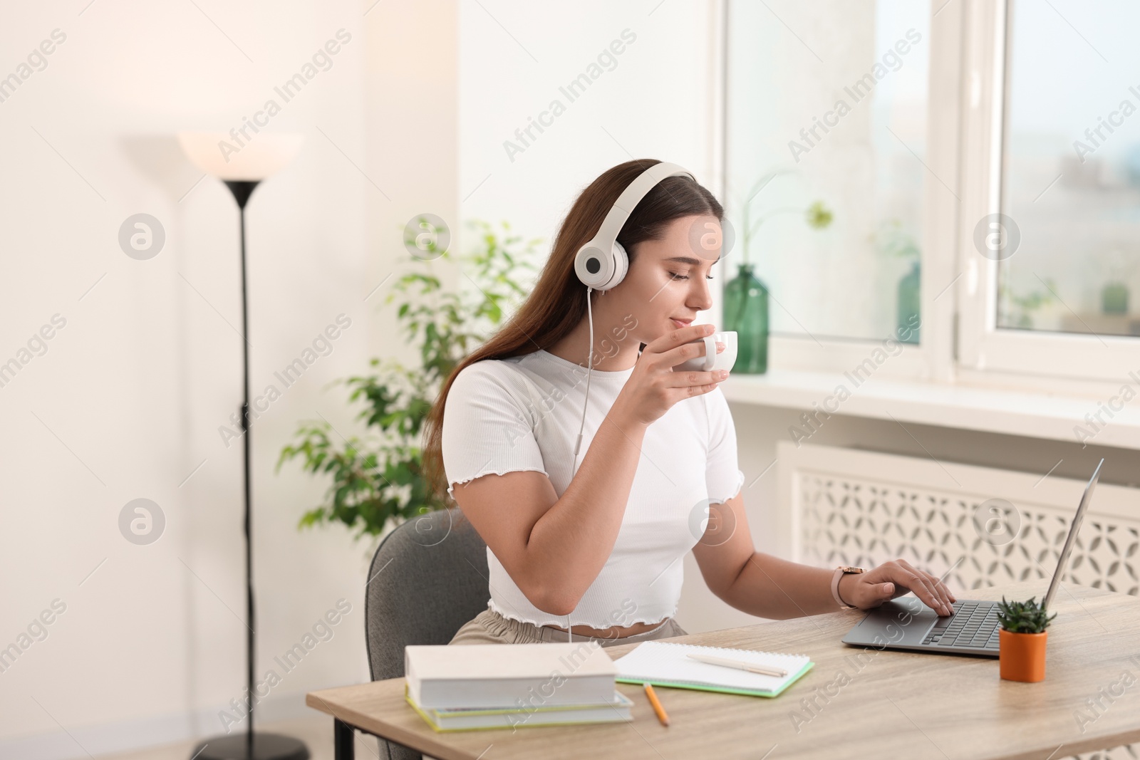 Photo of Student in headphones enjoying hot drink while studying with laptop at wooden table indoors