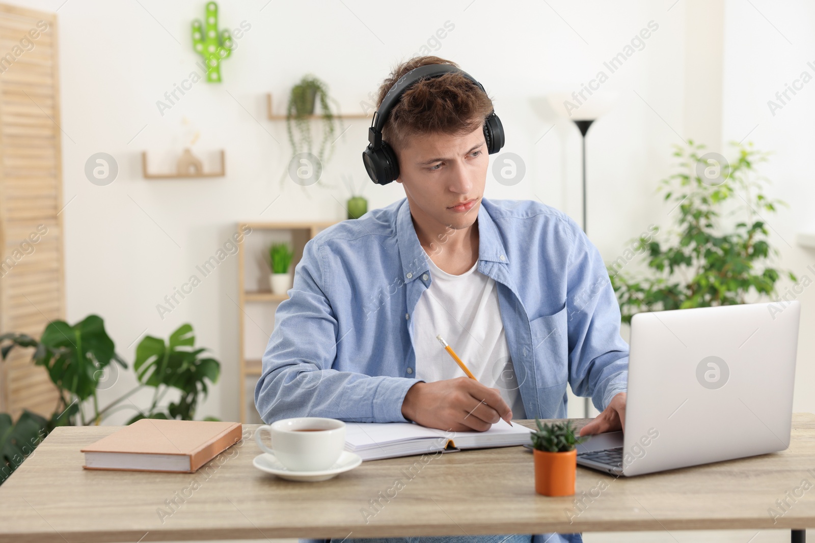 Photo of Student in headphones taking notes while studying with laptop at wooden table indoors