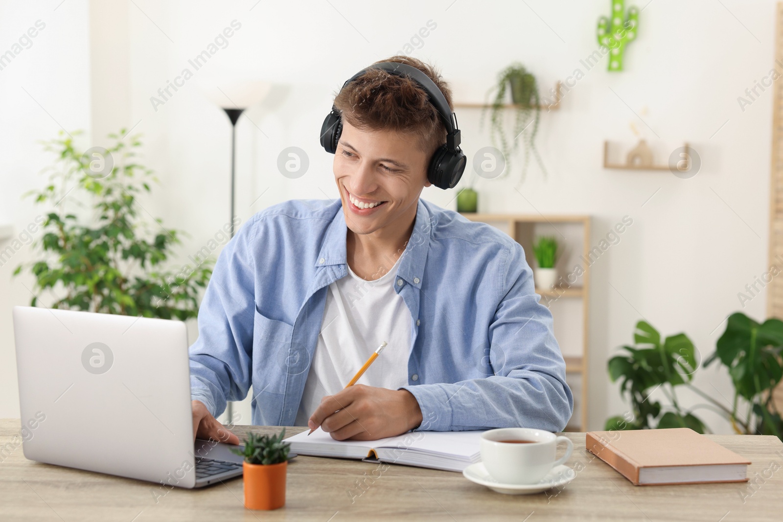 Photo of Student in headphones taking notes while studying with laptop at wooden table indoors