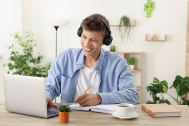 Photo of Student in headphones taking notes while studying with laptop at wooden table indoors