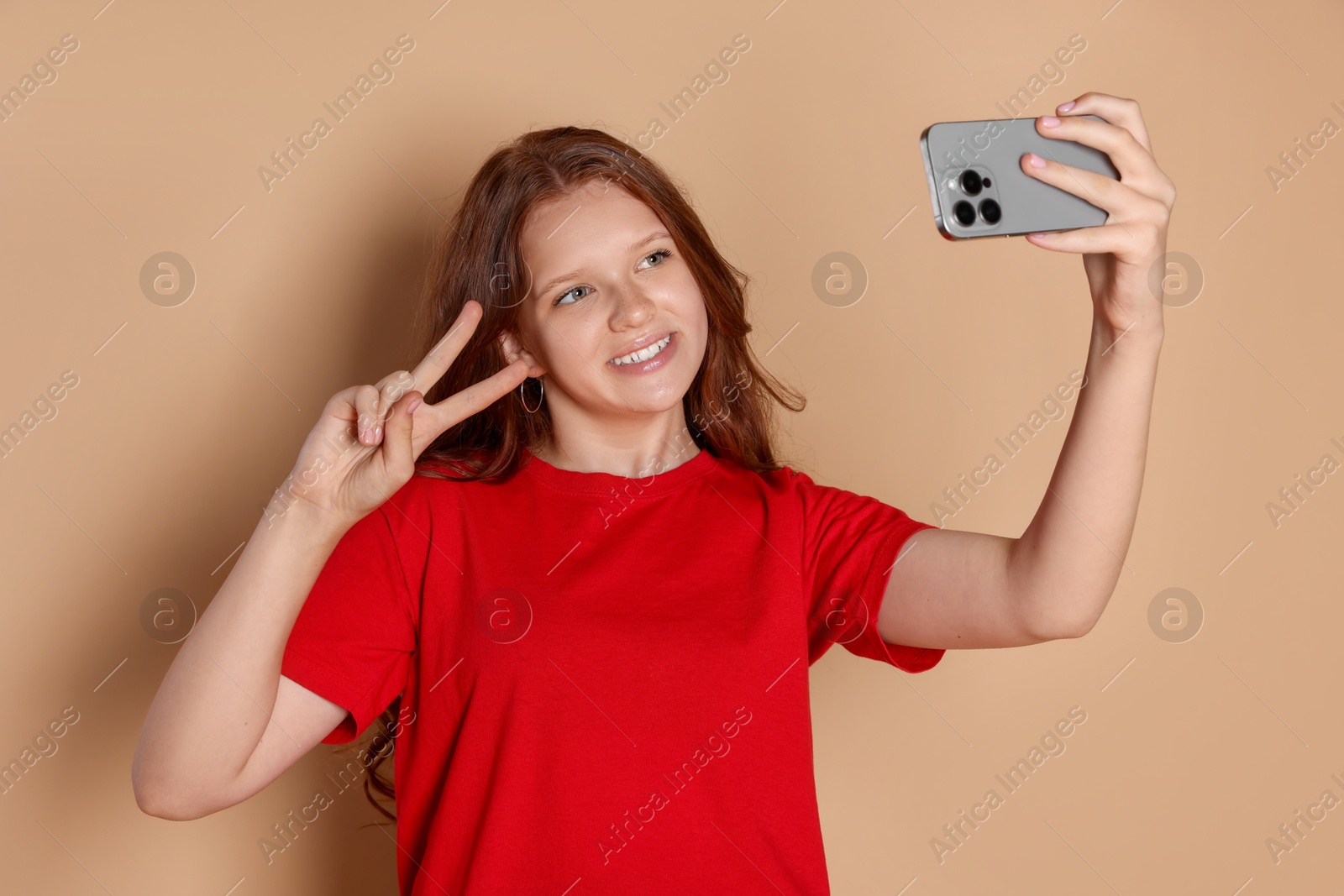 Photo of Smiling teenage girl taking selfie and showing peace sign on beige background