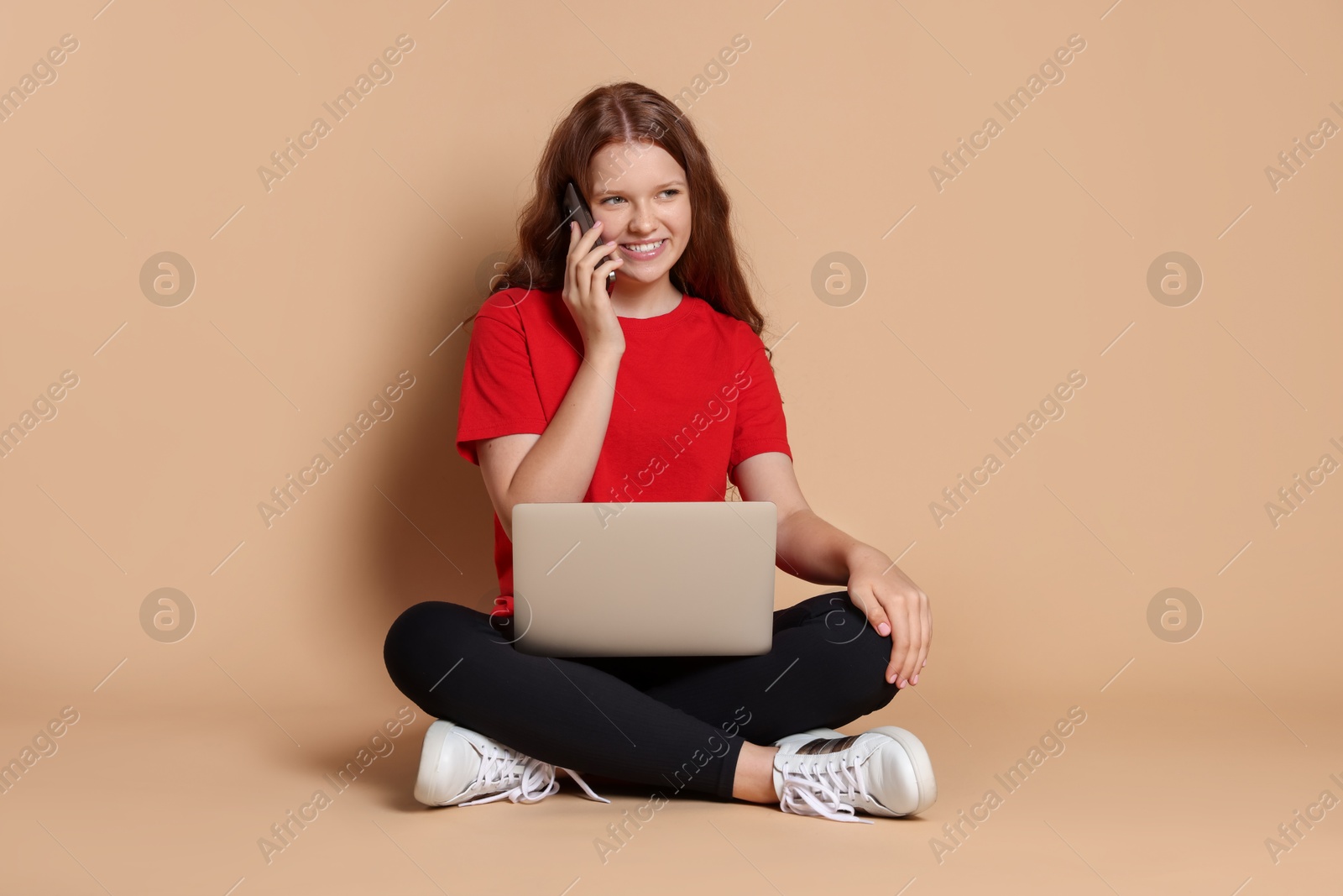 Photo of Smiling teenage girl with laptop talking by smartphone on beige background